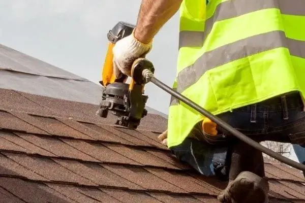Roofer in a high-visibility vest nailing brown asphalt shingles with a nail gun